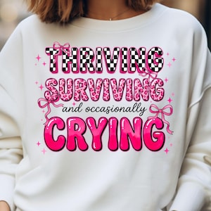 May include: White sweatshirt with the words "THRIVING, SURVIVING, and occasionally CRYING" in pink and black lettering. The word "THRIVING" is in a black and white checkered pattern. Pink bows and stars decorate the design.