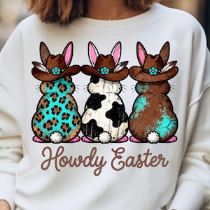 May include: White sweatshirt featuring three cartoon bunnies with cowboy hats. The bunnies have different patterns: teal leopard print, black and white cow print, and brown with teal accents. The text "Howdy Easter" is written in brown script.