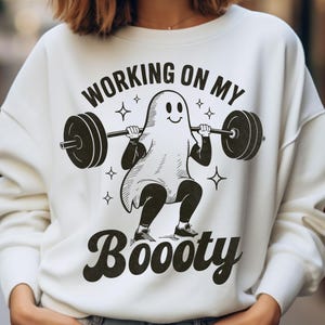 May include: White sweatshirt with a black graphic of a ghost lifting weights. The text "WORKING ON MY" is above the ghost, and "BOOOTY" is below. The design includes stars.