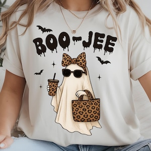 May include: Cream-colored t-shirt with a Halloween-themed graphic. The design features a ghost wearing sunglasses and a leopard print bow, holding a coffee cup and a tote bag. The text "BOO-JEE" is above the ghost, with bats and stars.