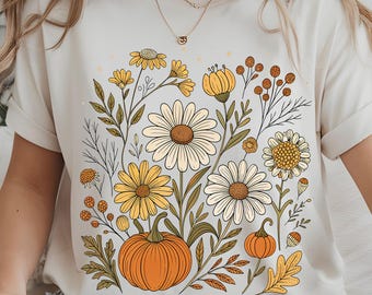 May include: Cream-colored t-shirt featuring a floral design with daisies, yellow flowers, and pumpkins. The illustration has a vintage aesthetic with brown, orange, and green accents. The shirt is a casual, everyday wear item.