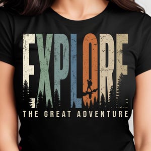 Puede incluir: Camiseta negra con la palabra "EXPLORE" en letras grandes, multicolores y desgastadas. El diseño incluye la silueta de un excursionista y árboles, con el texto "THE GREAT ADVENTURE" debajo.