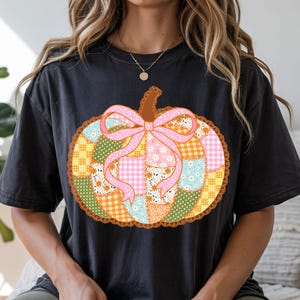 May include: Black t-shirt featuring a colorful patchwork pumpkin design. The pumpkin has a pink bow and a brown stem. The patchwork includes floral, gingham, and polka dot patterns in various colors. The design is outlined with a decorative orange lace.