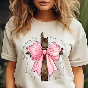 May include: A cream-coloured t-shirt with a graphic of a wooden cross and a large pink bow. The design includes the text "She is clothed with strength and dignity. She laughs without fear of the future. Proverbs 31:25."