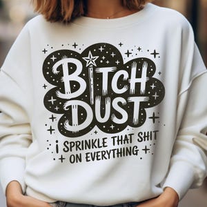 Puede incluir: Sudadera blanca con un diseño gráfico negro. El diseño presenta las palabras "BITCH DUST" en forma de nube, con una estrella y una varita, y la frase "I SPRINKLE THAT SHIT ON EVERYTHING".