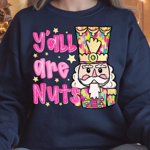 May include: Navy blue sweatshirt with the text "Y'all are Nuts" in pink, accompanied by star graphics. A colorful nutcracker graphic is on the right side, featuring a gold crown and a white beard.