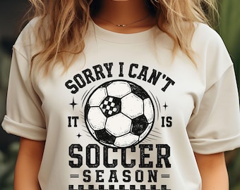 May include: A light beige t-shirt with the text "SORRY I CAN'T IT IS SOCCER SEASON" in black. The design includes a soccer ball graphic. The shirt has short sleeves and a crew neck.