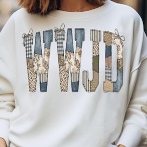 May include: White sweatshirt featuring the letters "WWJD" in a patchwork design. The letters are composed of various patterns, including floral, leopard print, and plaid, with small bow accents at the top of the "W" and "J" letters.