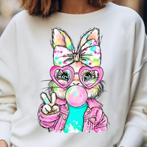 May include: White sweatshirt featuring a colorful cartoon bunny wearing heart-shaped glasses and a pink jacket, blowing a bubblegum bubble. The bunny has a large bow and is giving a peace sign.