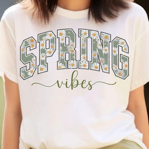 May include: White t-shirt with the words "SPRING" in a green and white floral design. The word "vibes" is written in a cursive font below the word "SPRING".