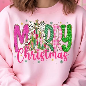 May include: Pink sweatshirt with a colorful "Merry Christmas" graphic. The graphic features a Christmas tree with a polka dot bow and the words "Merry Christmas" in pink, green, and plaid patterns. Small star and snowflake accents are scattered around the graphic.