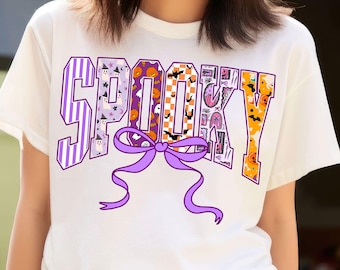 May include: White t-shirt with the word "SPOOKY" in purple and orange block letters. Each letter has a different Halloween-themed pattern, including stripes, bats, and pumpkins. A purple bow is centered below the word.