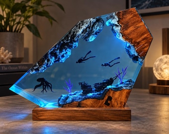 Puede incluir: Una lámpara decorativa con un bloque inclinado de madera y resina azul. Dentro de la resina hay siluetas de dos buceadores, un pulpo y un naufragio, iluminados por una luz azul interna. La pieza descansa sobre una base rectangular de madera.