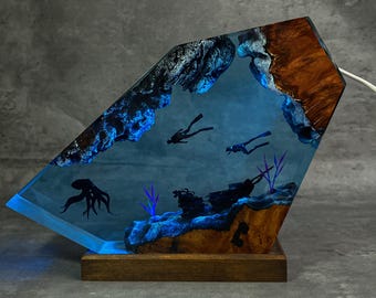 May include: A unique, illuminated resin lamp featuring an underwater scene. The lamp showcases a blue ocean with divers, an octopus, and a shipwreck. The base is wood, and the lamp has a geometric shape.