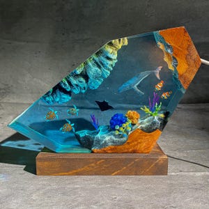 May include: A decorative resin lamp featuring an underwater scene. The lamp showcases a whale, turtles, and colorful coral within a clear, angled resin block. The base is made of dark wood, and the lamp has a warm, inviting glow.