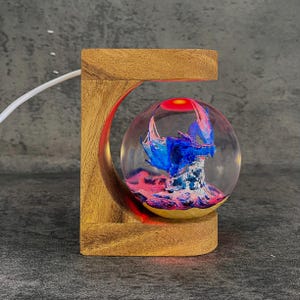 May include: A decorative wooden base with a clear orb containing a blue and pink sculpture. The orb is lit with a red light at the top. The wooden base has a curved design and a natural wood finish.