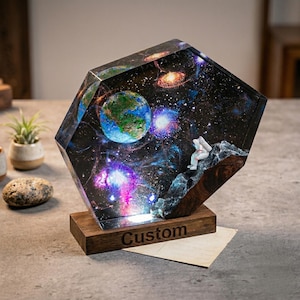 May include: A decorative resin sculpture featuring a detailed depiction of space, including planets, galaxies, and an astronaut. The sculpture is mounted on a wooden base with the word "Custom" engraved on it. The artwork showcases vibrant colors and intricate details.