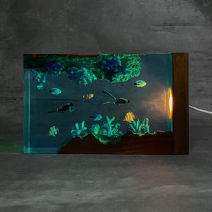 May include: A rectangular resin lamp featuring an underwater scene with coral, fish, and divers. The lamp has a wooden frame and an illuminated section on the side. The scene includes various colorful fish and coral formations.