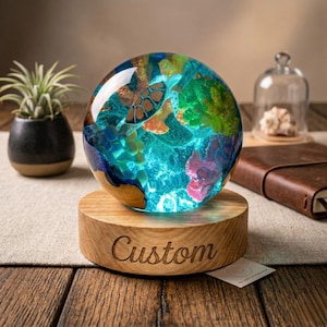 May include: A decorative glass sphere with a sea turtle design, set on a wooden base engraved with the word "Custom". The sphere features a vibrant underwater scene with coral and a turtle, illuminated with blue light.