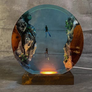 May include: A round resin lamp featuring a miniature underwater scene with two divers. The lamp has a wooden base and a clear resin dome with a blue background. The scene includes rock formations and a warm, glowing light at the bottom. The lamp is approximately 8 inches in diameter.