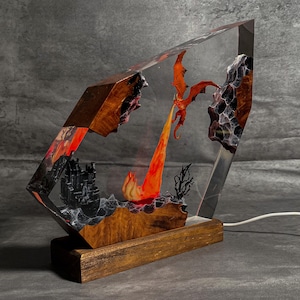 May include: A fantasy-themed resin lamp featuring a fiery orange dragon breathing fire. The lamp has a wooden base and a clear, angled resin body with a castle and rock formations. The dragon is in mid-flight, with the fire illuminating the scene.