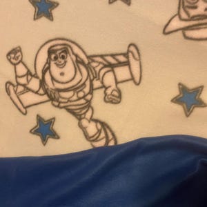 May include: A white fleece blanket with a blue and white pattern of stars and the character Buzz Lightyear from the Toy Story movie franchise. The blanket is folded in half and the bottom half is a solid blue color.