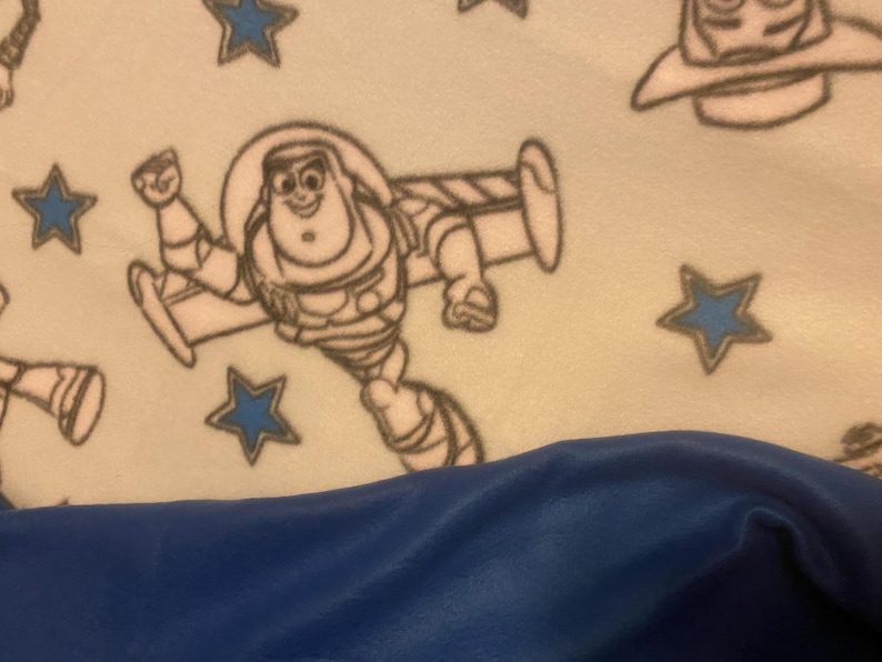 May include: A white fleece blanket with a blue and white pattern of stars and the character Buzz Lightyear from the Toy Story movie franchise. The blanket is folded in half and the bottom half is a solid blue color.
