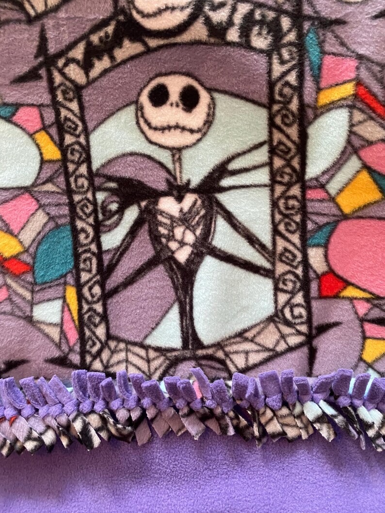 Puede incluir: Una manta de forro polar morada con un dise&ntilde;o de vidriera de colores que presenta a Jack Skellington de Pesadilla antes de Navidad. La manta tiene un borde de flecos morados.
