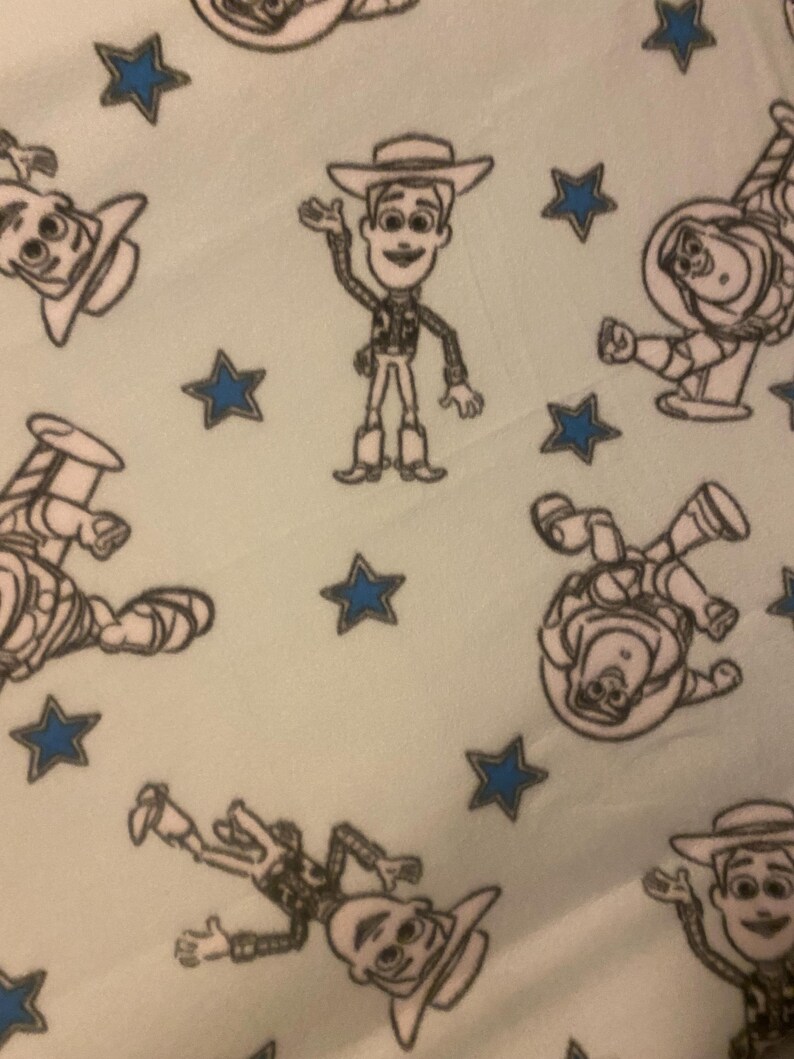 May include: A light blue fabric with a repeating pattern of black and white cartoon characters from the Toy Story movie. The characters include Woody, Buzz Lightyear, and a few stars.