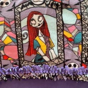 Puede incluir: Manta de forro polar morada con un estampado de Pesadilla Antes de Navidad con Sally y Jack Skellington. La manta tiene un ribete de flecos morados.