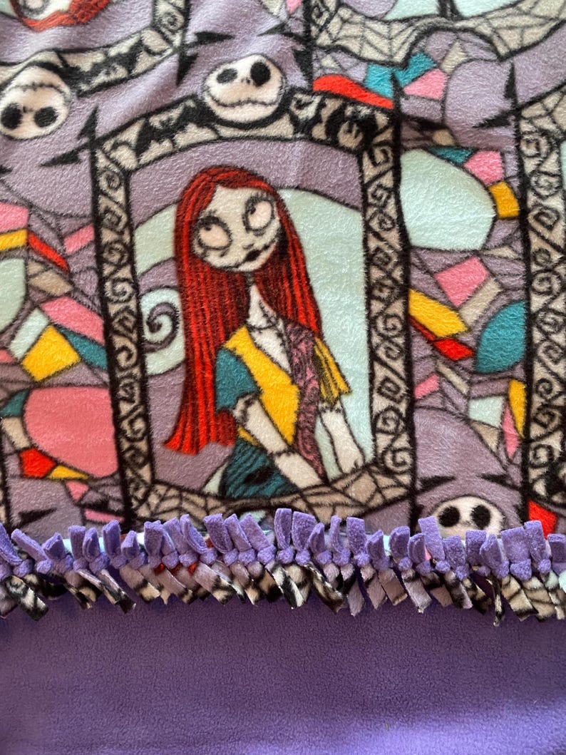 Puede incluir: Manta de forro polar morada con un estampado de Pesadilla Antes de Navidad con Sally y Jack Skellington. La manta tiene un ribete de flecos morados.