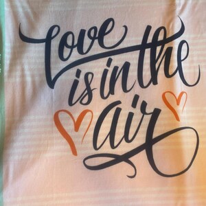 May include: A pink and white striped fabric with the phrase "Love is in the air" in black script. Two orange hearts are below the word "air". The fabric has a light green border.