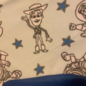 May include: A white fleece blanket with a repeating pattern of blue stars and cartoon images of Woody from Toy Story. The blanket is folded in half and a blue surface is visible in the bottom of the image.