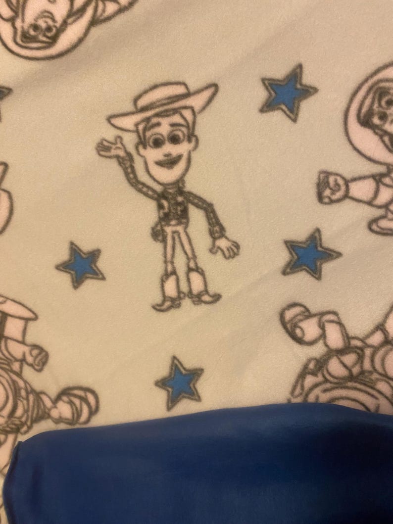 May include: A white fleece blanket with a repeating pattern of blue stars and cartoon images of Woody from Toy Story. The blanket is folded in half and a blue surface is visible in the bottom of the image.
