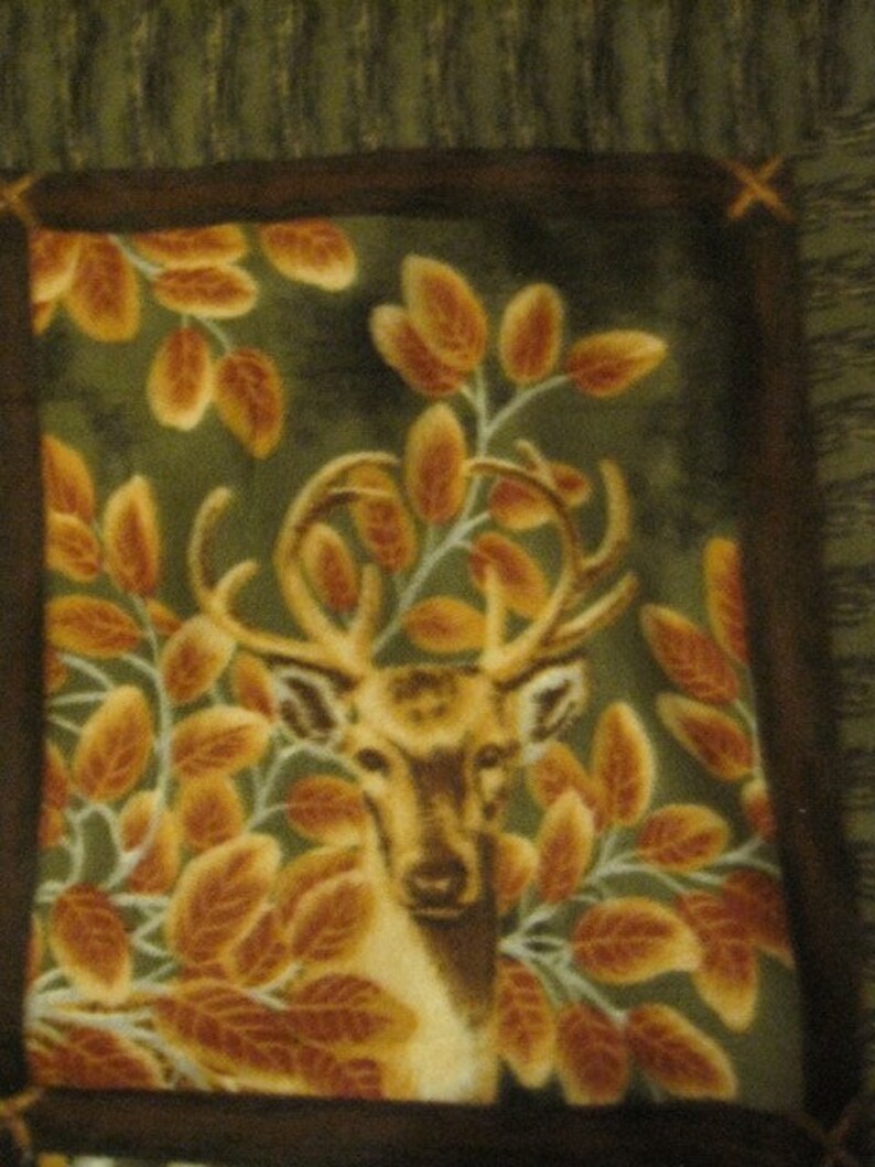 Pode incluir: Um veado marrom e branco com chifres est&aacute; rodeado de folhas laranja e marrons sobre um fundo verde. O veado est&aacute; olhando diretamente para o espectador. A imagem &eacute; emoldurada com uma borda marrom.