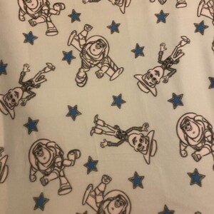May include: A light blue fabric with a repeating pattern of white cartoon characters and blue stars. The characters are of a space ranger and a cowboy.