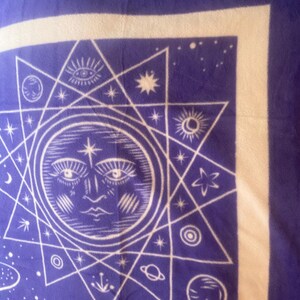 May include: A purple tapestry with a white sun and stars design. The sun has a face and is surrounded by a starburst pattern. The tapestry has a white border.