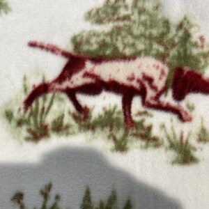 May include: A white fabric with a brown and white dog running through green bushes. The dog is a hunting breed with long ears and a long tail.