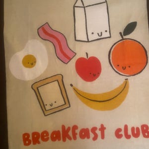 May include: A beige tote bag featuring a whimsical breakfast-themed design. The illustration includes a fried egg, bacon, milk carton, apple, orange, toast, and banana, all with smiling faces. The words "BREAKFAST CLUB" are printed in red.