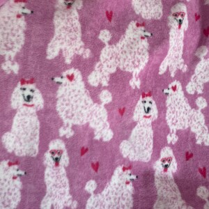 May include: Pink fleece fabric with a repeating pattern of white poodles wearing red bows and sunglasses.  Pink hearts are scattered throughout the pattern.