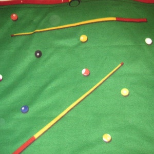 May include: A green felt pool table with two cues, one yellow and one red, and several colorful billiard balls scattered across the surface.