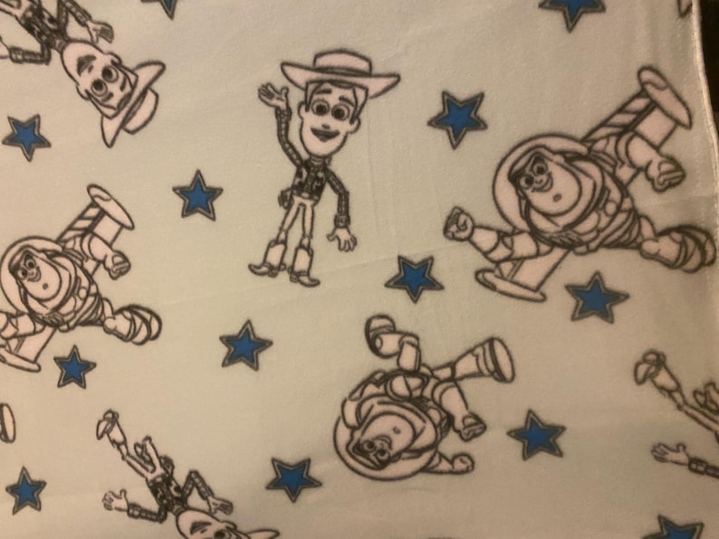 May include: A light blue fabric with a repeating pattern of Woody and Buzz Lightyear from Toy Story. The characters are outlined in black and the background is a light blue color. There are also blue stars scattered throughout the fabric.