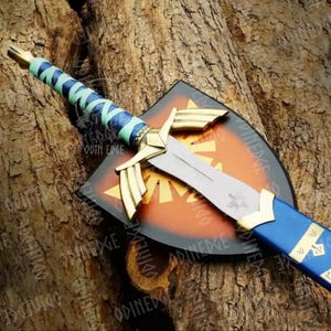 May include: A decorative sword and shield set. The sword has a silver blade, gold details, and a blue and green wrapped handle. The shield is black with a red and gold emblem. The set is displayed against a textured wooden background.