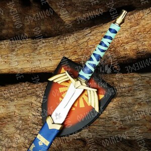 May include: A decorative sword and shield set. The sword has a silver blade, gold details, and a blue and teal wrapped handle. The shield is black with a red and gold design. The set is displayed on a wooden surface.