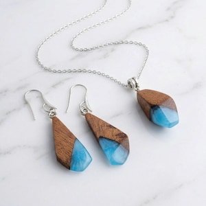 Op de afbeelding: Een sieradenset met een hangerketting en bijpassende oorbellen. De sieraden hebben een zilveren ketting en haken. De hanger en oorbellen hebben een houten bovenkant en een blauwe harsbodem. De set wordt gepresenteerd op een wit marmeren oppervlak.