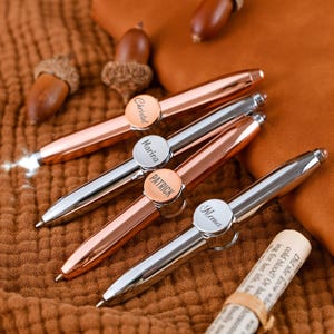 May include: Four personalized pens with names engraved on circular accents. Two pens are silver, and two are rose gold. The pens are arranged on a textured brown surface, with acorns and a rolled-up paper scroll nearby.