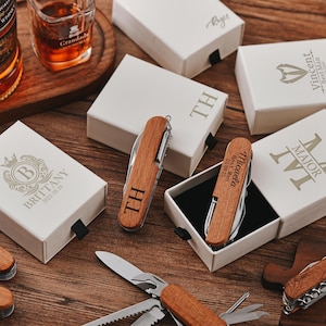 May include: Wooden pocket knives with personalized engravings, displayed with gift boxes. The knives have silver blades and are engraved with initials and names. The boxes are white with custom text and logos.