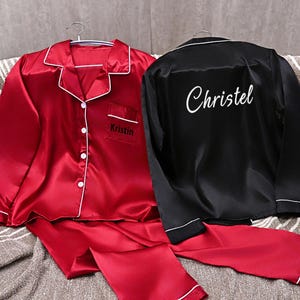 May include: Two sets of satin pajamas. One set is red with white trim and the name "Kristin" embroidered on the pocket. The other set is black with white trim and the name "Christel" embroidered on the back. Both sets are displayed on hangers.