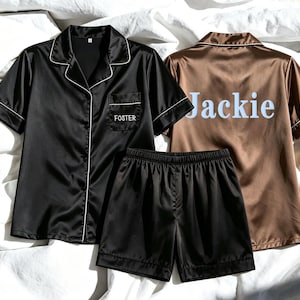 May include: Black and brown satin pajama sets with white trim are displayed on a white sheet. One black set has "Foster" embroidered on the pocket. The brown set has "Jackie" printed on the back in light blue.