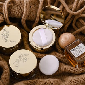 May include: An assortment of beauty products, including two gold compacts with floral designs and the name "Jenny", a round wooden object, a perfume bottle with the word "FREEDOM", and a white powder puff. The items are arranged on a brown knitted surface.